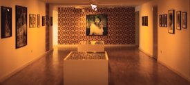 A gallery space featuring artwork on both side walls and a large framed portrait in the center on the back wall. The room has a warm, dim lighting with a patterned wallpaper as a backdrop. Display pedestals are situated in the foreground.