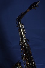 A brass saxophone with intricate keys and levers is set against a deep blue background. The polished metal surface reflects light, highlighting the instrument's craftsmanship.