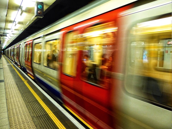 A subway train moves quickly through an underground station, causing a blur effect. The platform is empty, lined with safety markers and overhead lights. The train has a red and white color scheme, with some passengers visible through the windows. A green signal light is visible above the train.