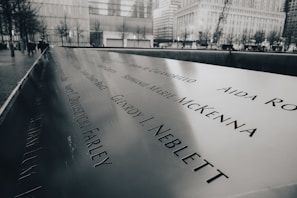 A memorial with engraved names on a polished metal surface, set in an urban environment. The background features tall buildings and bare trees, suggesting a city landscape. The atmosphere is somber and reflective, with a monochrome color palette.