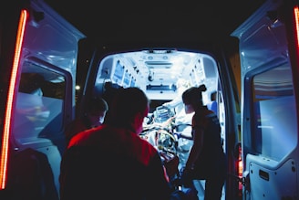 An ambulance is parked with its rear doors open, revealing a brightly lit interior. Several medical professionals are working intently inside, attending to a patient on a stretcher. The scene is illuminated by the cool blue and white lights of the ambulance, contrasting with the dark surroundings.