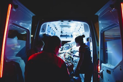 An ambulance is parked with its rear doors open, revealing a brightly lit interior. Several medical professionals are working intently inside, attending to a patient on a stretcher. The scene is illuminated by the cool blue and white lights of the ambulance, contrasting with the dark surroundings.