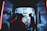 An ambulance is parked with its rear doors open, revealing a brightly lit interior. Several medical professionals are working intently inside, attending to a patient on a stretcher. The scene is illuminated by the cool blue and white lights of the ambulance, contrasting with the dark surroundings.