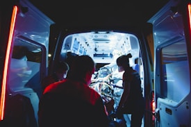 An ambulance is parked with its rear doors open, revealing a brightly lit interior. Several medical professionals are working intently inside, attending to a patient on a stretcher. The scene is illuminated by the cool blue and white lights of the ambulance, contrasting with the dark surroundings.