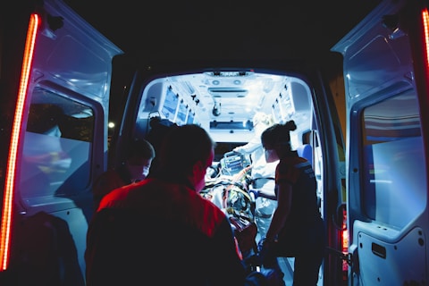 An ambulance is parked with its rear doors open, revealing a brightly lit interior. Several medical professionals are working intently inside, attending to a patient on a stretcher. The scene is illuminated by the cool blue and white lights of the ambulance, contrasting with the dark surroundings.
