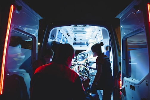 An ambulance is parked with its rear doors open, revealing a brightly lit interior. Several medical professionals are working intently inside, attending to a patient on a stretcher. The scene is illuminated by the cool blue and white lights of the ambulance, contrasting with the dark surroundings.