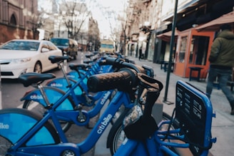 A row of blue rental bicycles is lined up on a city street. The bicycles are branded with 'citi bike' and have black handlebars. In the background, there are buildings with storefronts, a person in a jacket walking, and several stationary cars, including a white sedan.