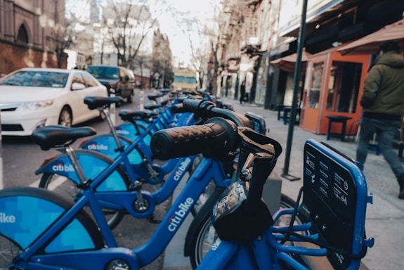 A row of blue rental bicycles is lined up on a city street. The bicycles are branded with 'citi bike' and have black handlebars. In the background, there are buildings with storefronts, a person in a jacket walking, and several stationary cars, including a white sedan.