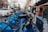 A row of blue rental bicycles is lined up on a city street. The bicycles are branded with 'citi bike' and have black handlebars. In the background, there are buildings with storefronts, a person in a jacket walking, and several stationary cars, including a white sedan.
