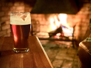 A pint of beer sits on a wooden table in a warm, cozy setting with a fire burning in the background. The flames illuminate the room, casting a soft glow on the beer and creating a comforting atmosphere.
