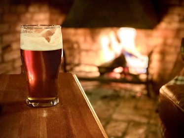 A pint of beer sits on a wooden table in a warm, cozy setting with a fire burning in the background. The flames illuminate the room, casting a soft glow on the beer and creating a comforting atmosphere.