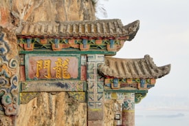 An ornate, ancient-style architectural structure with vibrant colors and intricate designs. It features traditional curved roof tiles and decorative carvings, with prominent Chinese characters in gold on a central panel. The background has a rocky texture and a hazy view of a distant landscape.