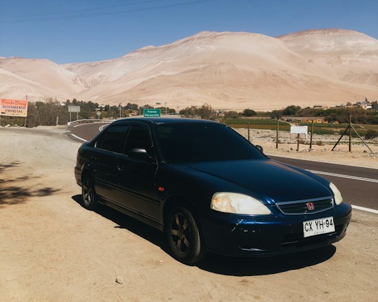 A dark-colored car is parked beside a road with a mountainous desert landscape in the background. The road signs indicate nearby points of interest. The sky is clear, suggesting a sunny day.