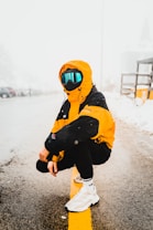 A person dressed in a bright orange and black winter jacket with a hood and reflective ski goggles crouches on a snow-lined road. The background is snowy, with blurred cars and structures barely visible. The person wears black pants and white sneakers, suggesting a mix of sports and street style wear.