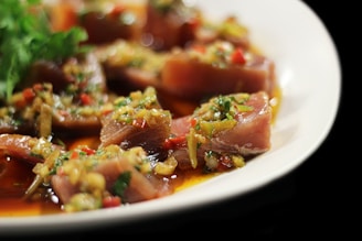 Sliced pieces of tuna, possibly raw, topped with a colorful mix of chopped vegetables and herbs, including red peppers and green parsley, all drizzled with a rich sauce. The dish is presented on a white plate with a glossy shine indicating freshness.