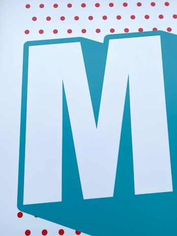 A large, bold, white letter 'M' is centered against a teal blue background. The letter is outlined with a thicker shade of teal that matches the background. Above and below the letter are multiple small, evenly spaced red dots on a white background.