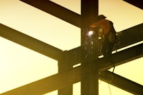 A construction worker is welding on a steel framework high above the ground. Sparks fly from the welding point, and the worker is wearing safety gear including a helmet and harness. The scene is backlit by a soft, warm yellow light.