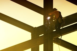 A construction worker is welding on a steel framework high above the ground. Sparks fly from the welding point, and the worker is wearing safety gear including a helmet and harness. The scene is backlit by a soft, warm yellow light.