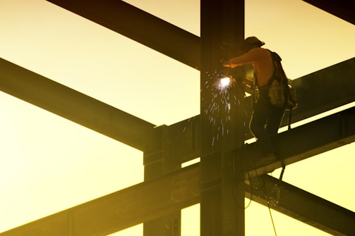 A construction worker is welding on a steel framework high above the ground. Sparks fly from the welding point, and the worker is wearing safety gear including a helmet and harness. The scene is backlit by a soft, warm yellow light.
