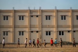A group of people engage in a casual volleyball game on a grassy field with a backdrop of a beige apartment building featuring multiple windows. The individuals are dressed in casual attire and appear focused on the game, with a volleyball visible in mid-air.