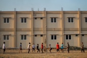 A group of people engage in a casual volleyball game on a grassy field with a backdrop of a beige apartment building featuring multiple windows. The individuals are dressed in casual attire and appear focused on the game, with a volleyball visible in mid-air.