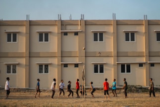A group of people engage in a casual volleyball game on a grassy field with a backdrop of a beige apartment building featuring multiple windows. The individuals are dressed in casual attire and appear focused on the game, with a volleyball visible in mid-air.