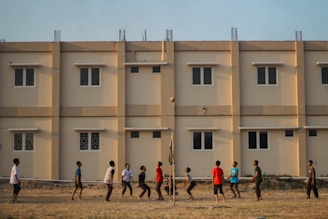A group of people engage in a casual volleyball game on a grassy field with a backdrop of a beige apartment building featuring multiple windows. The individuals are dressed in casual attire and appear focused on the game, with a volleyball visible in mid-air.