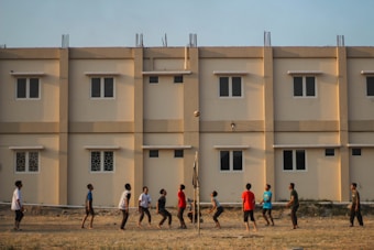 A group of people engage in a casual volleyball game on a grassy field with a backdrop of a beige apartment building featuring multiple windows. The individuals are dressed in casual attire and appear focused on the game, with a volleyball visible in mid-air.