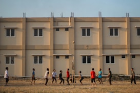 A group of people engage in a casual volleyball game on a grassy field with a backdrop of a beige apartment building featuring multiple windows. The individuals are dressed in casual attire and appear focused on the game, with a volleyball visible in mid-air.