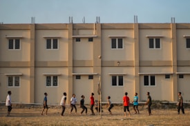 A group of people engage in a casual volleyball game on a grassy field with a backdrop of a beige apartment building featuring multiple windows. The individuals are dressed in casual attire and appear focused on the game, with a volleyball visible in mid-air.