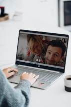 A person is using a laptop, which displays a video call with several smiling individuals on the screen. Their hands are on the keyboard, and there is a cup of coffee nearby.