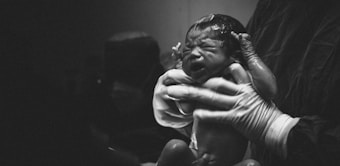 A newborn baby being held by a person wearing gloves and a protective gown, possibly a medical professional. The setting appears to be a hospital or delivery room. The baby is wet and crying, indicating it has just been delivered.
