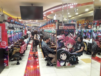 A bustling arcade filled with people playing various gaming machines. Brightly colored displays and animated characters are prominent on posters and screens. The atmosphere is lively with patrons seated in rows, focusing on the games. Overhead signage and electronic scoreboards are visible, contributing to the busy and vibrant environment.