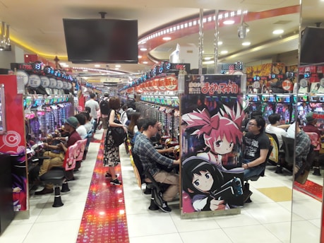 A bustling arcade filled with people playing various gaming machines. Brightly colored displays and animated characters are prominent on posters and screens. The atmosphere is lively with patrons seated in rows, focusing on the games. Overhead signage and electronic scoreboards are visible, contributing to the busy and vibrant environment.