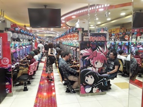 A bustling arcade filled with people playing various gaming machines. Brightly colored displays and animated characters are prominent on posters and screens. The atmosphere is lively with patrons seated in rows, focusing on the games. Overhead signage and electronic scoreboards are visible, contributing to the busy and vibrant environment.