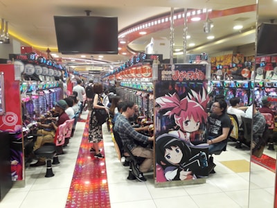 A bustling arcade filled with people playing various gaming machines. Brightly colored displays and animated characters are prominent on posters and screens. The atmosphere is lively with patrons seated in rows, focusing on the games. Overhead signage and electronic scoreboards are visible, contributing to the busy and vibrant environment.