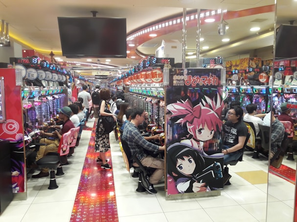 A bustling arcade filled with people playing various gaming machines. Brightly colored displays and animated characters are prominent on posters and screens. The atmosphere is lively with patrons seated in rows, focusing on the games. Overhead signage and electronic scoreboards are visible, contributing to the busy and vibrant environment.