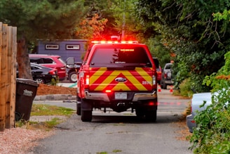 A red emergency vehicle is parked on a narrow road surrounded by greenery. The vehicle has flashing red lights and a distinctive yellow and red striped pattern on its rear. Several other vehicles are parked in the background, and orange traffic cones line the road. A trash bin is visible next to a wooden fence on the left.