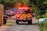 A red emergency vehicle is parked on a narrow road surrounded by greenery. The vehicle has flashing red lights and a distinctive yellow and red striped pattern on its rear. Several other vehicles are parked in the background, and orange traffic cones line the road. A trash bin is visible next to a wooden fence on the left.