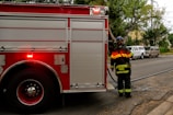 A firefighter in uniform stands near a large red fire truck, which is parked on a street. The fire truck has rolled-up doors, and there is a hose visible extending from it. Nearby, there are several parked vehicles, including two vans. Trees line the sidewalk, and a house can be seen in the background.