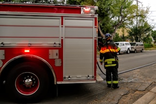 A firefighter in uniform stands near a large red fire truck, which is parked on a street. The fire truck has rolled-up doors, and there is a hose visible extending from it. Nearby, there are several parked vehicles, including two vans. Trees line the sidewalk, and a house can be seen in the background.