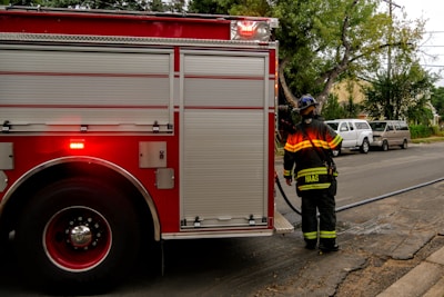 A firefighter in uniform stands near a large red fire truck, which is parked on a street. The fire truck has rolled-up doors, and there is a hose visible extending from it. Nearby, there are several parked vehicles, including two vans. Trees line the sidewalk, and a house can be seen in the background.