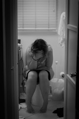 A person sitting on a toilet in a bathroom with their head in their hands, conveying a sense of distress or contemplation. The room has closed blinds, a trash bin, and various toiletries scattered around.