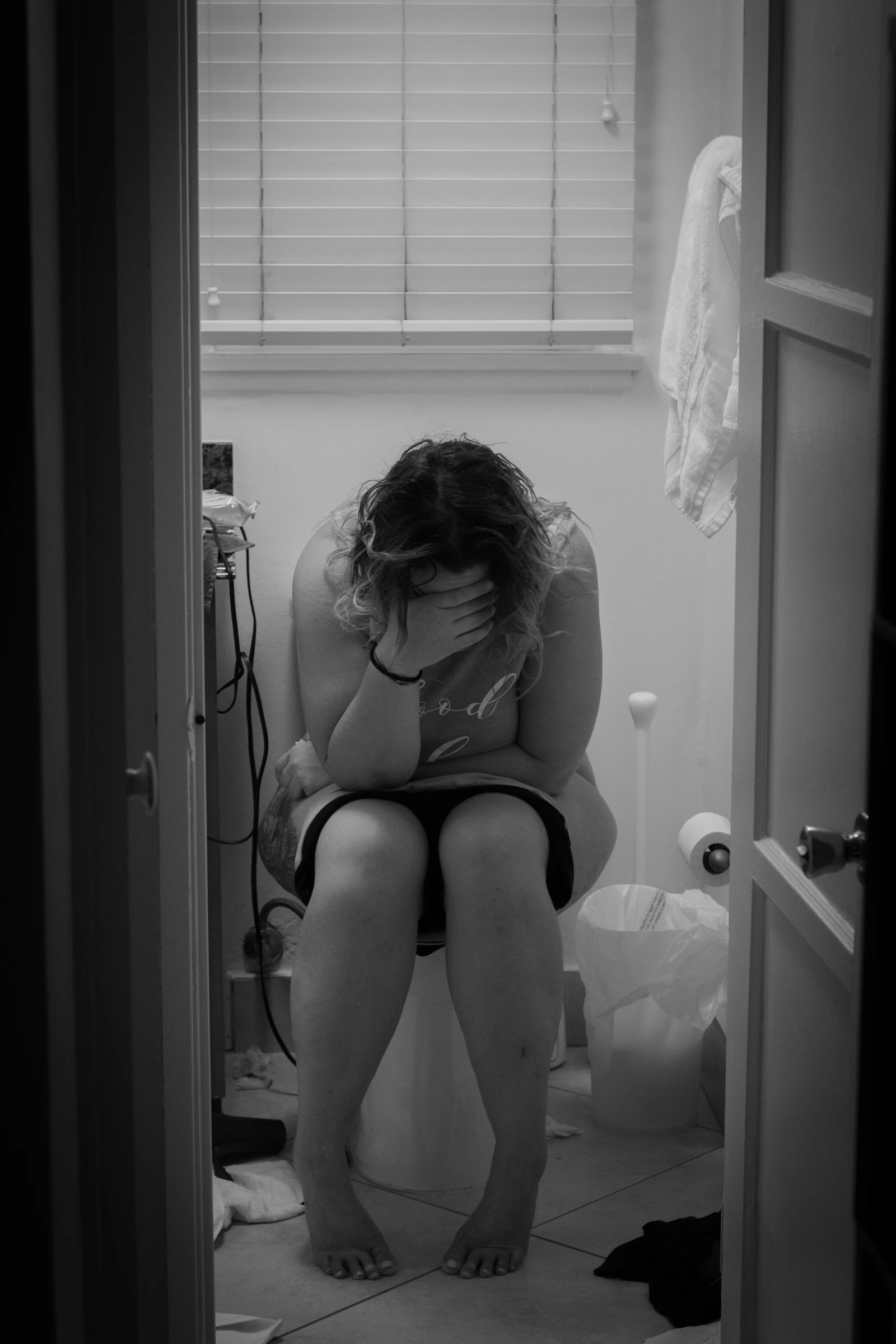 A new mother in a white tank top sitting on a toilet in the early postpartum recovery days
