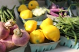 A variety of fresh vegetables including yellow bell peppers, green beans, and purple turnips are arranged in teal cartons, displayed on a light surface in natural lighting.
