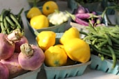 A variety of fresh vegetables including yellow bell peppers, green beans, and purple turnips are arranged in teal cartons, displayed on a light surface in natural lighting.