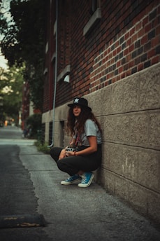 A person with long hair wearing a black cap and holding a camera sits against a brick wall on a narrow street. They are dressed casually in a gray t-shirt, black pants, and blue sneakers. The environment suggests an urban setting with trees visible in the background along the alley.