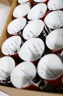 Several paper cups with handwritten text in Arabic are arranged in rows. The cups have a red exterior and white interior with black decorative patterns.