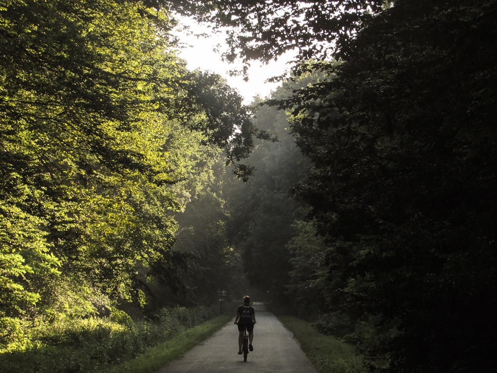 Hombre con chaqueta negra y pantalones negros caminando por el camino entre árboles verdes durante el día