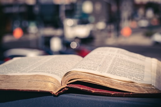 An open book with visible text pages, placed against a softly focused urban background. The book's pages appear slightly aged, and the background suggests a cityscape with blurred details of buildings and street features.